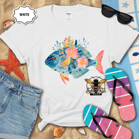 White t-shirt with a colorful fish design, surrounded by beach accessories on a sandy background.