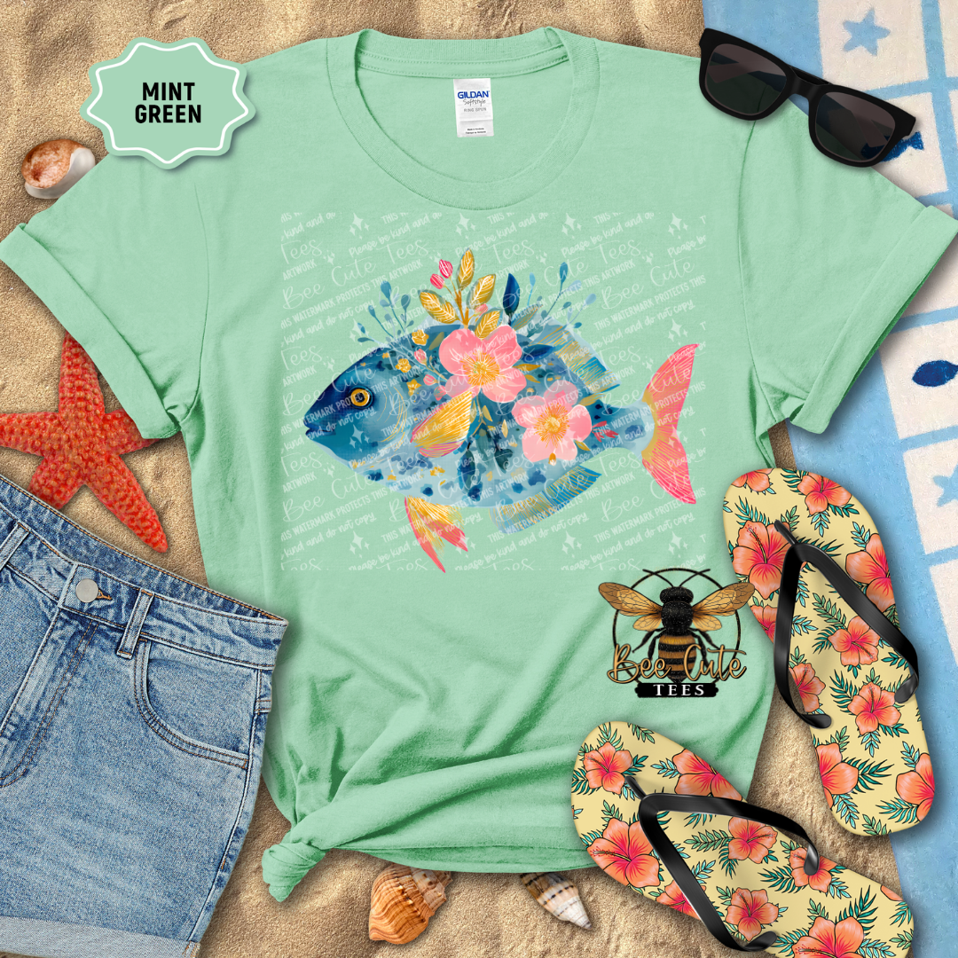 Mint green t-shirt with a colorful fish design, surrounded by beach accessories on a sandy background.