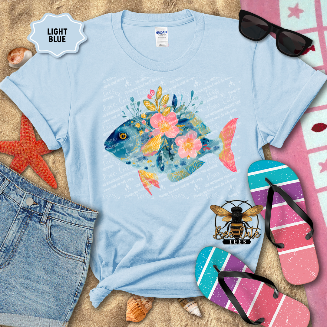 Light blue t-shirt with a colorful fish design on a sandy background with beach accessories.