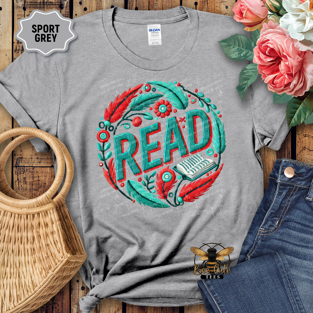 Gray t-shirt with 'READ' design on a wooden surface with flowers and a bag.