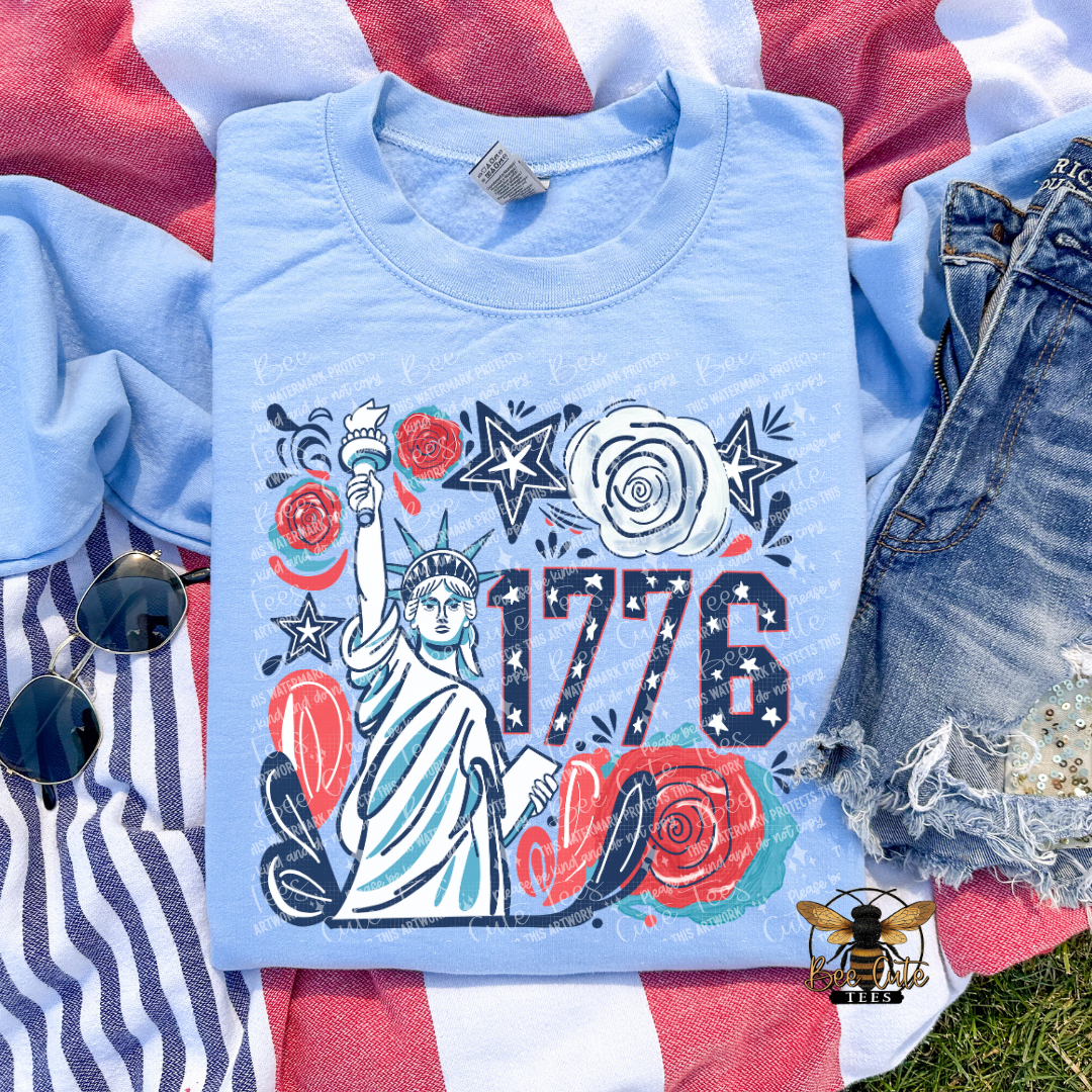 Light blue t-shirt with '1776' design featuring the Statue of Liberty on a flag background.