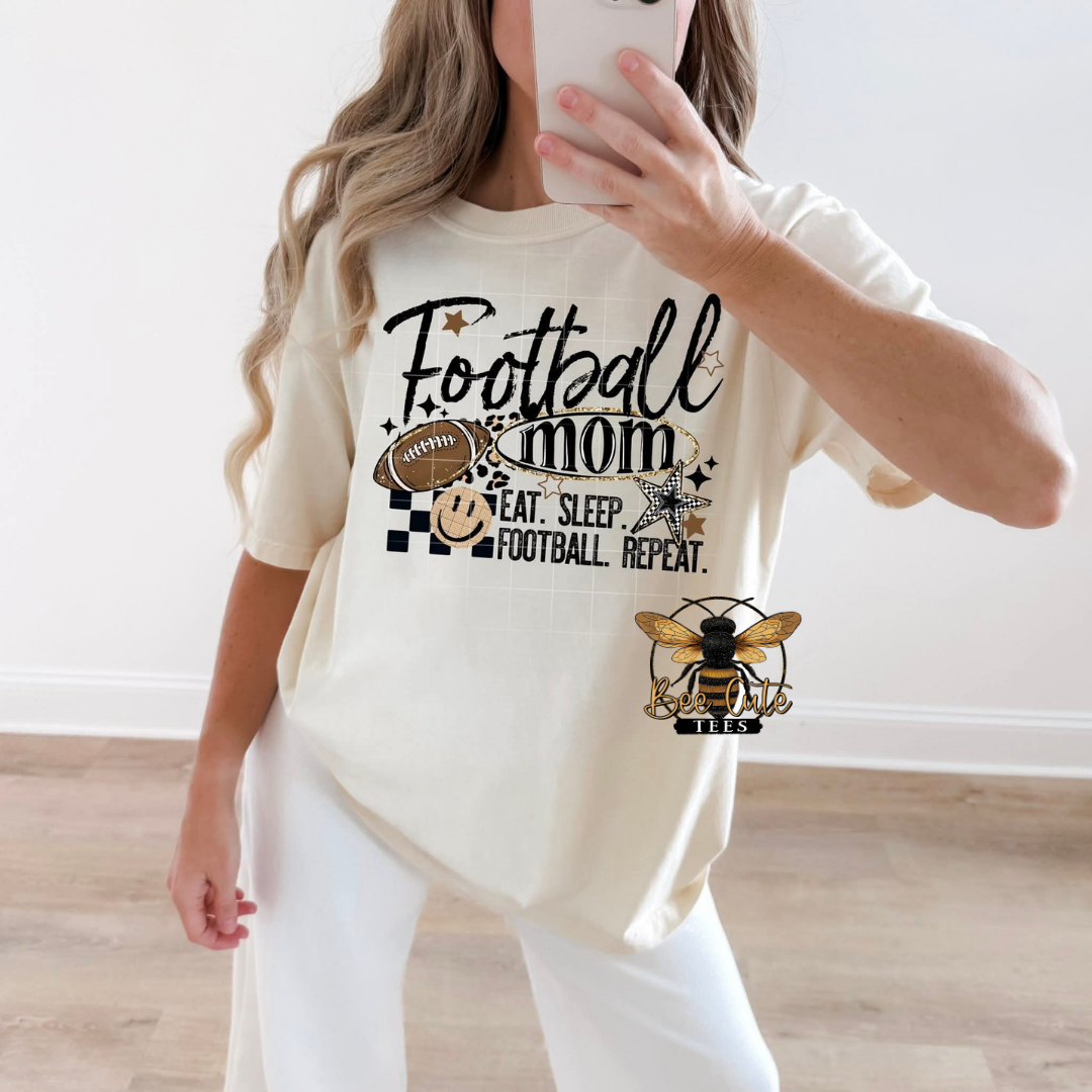 A woman takes a mirror selfie wearing a cream-colored graphic t-shirt with 'Football Mom' printed in black script, accompanied by a football and a bee logo with 'EAT. SLEEP.