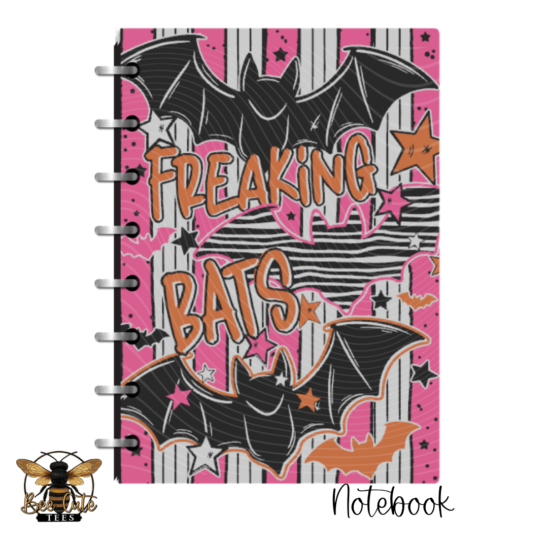 A spiral-bound notebook with a Halloween-themed cover featuring black bats, pink and white stripes, and the words 'FREAKING BATS' in orange. A bee logo with 'BEE'S' appears in the bottom-left corner, and 'Notebook' is written in script at the bottom-right.