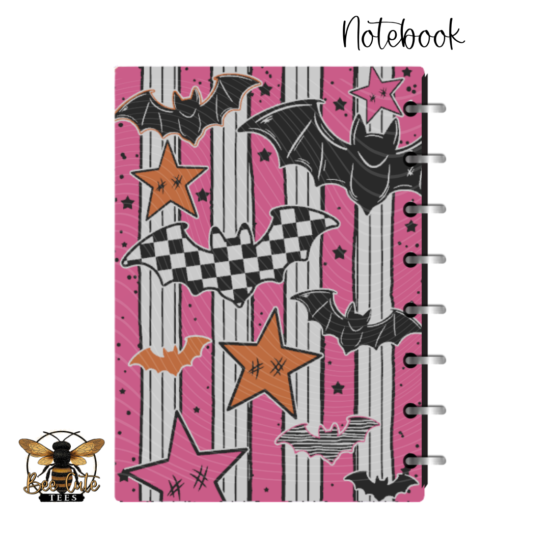 A spiral-bound notebook with a Halloween-themed cover featuring bats, stars, and stripes in pink, black, white, and orange, shown on a white background with a bee logo in the bottom-left corner and the word 'Notebook' at the top-right.