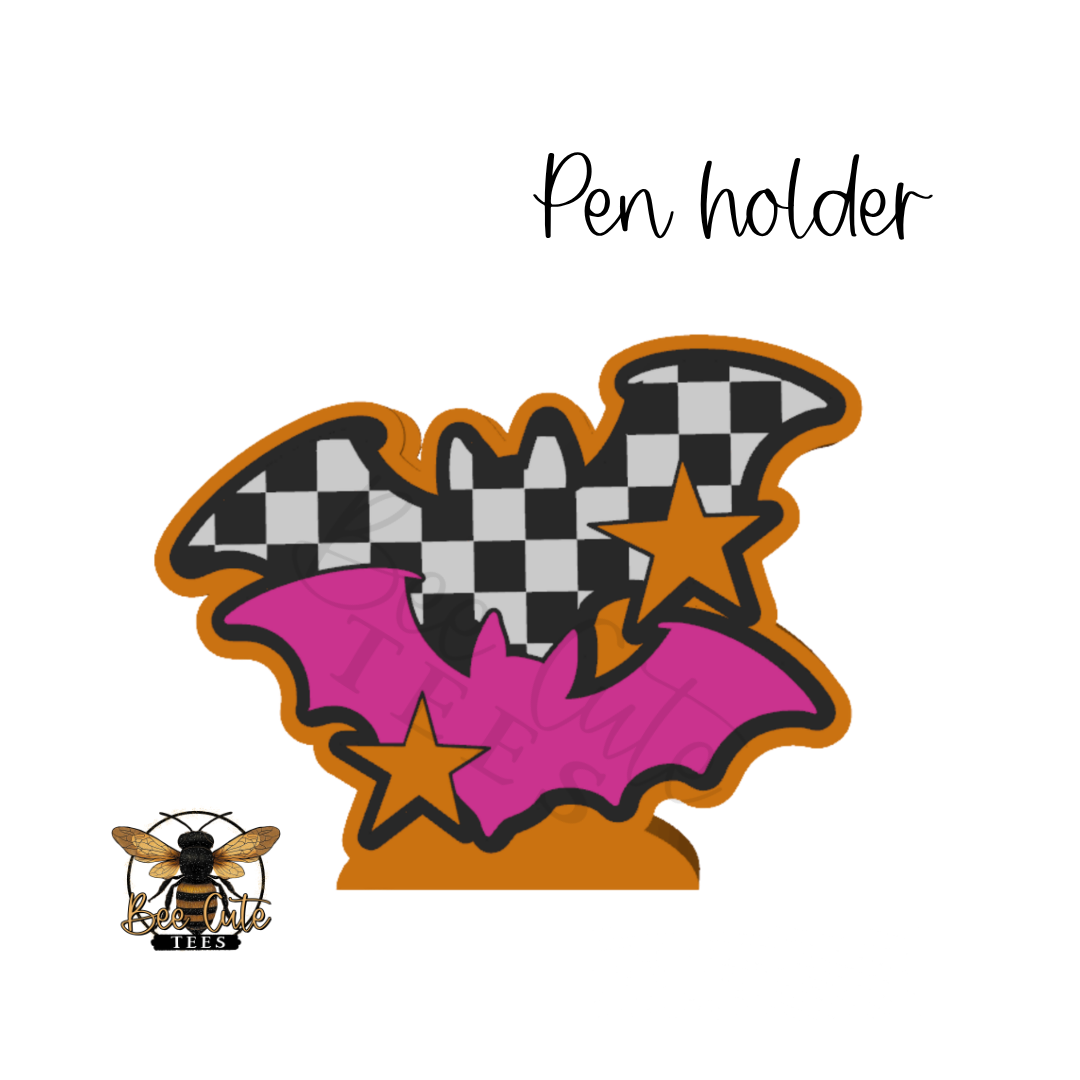 A Halloween-themed bat-shaped pen holder with a black and white checkered wing, pink wing, orange border, and gold stars, shown on a white background with the text 'Pen holder' and a bee logo.