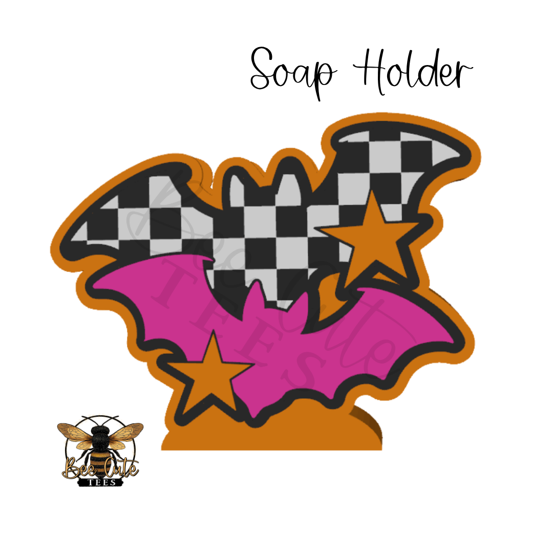 A Halloween-themed bat-shaped soap holder with a black and white checkered wing, bright pink wing, orange border, and orange stars, shown on a white background with the text 'Soap Holder' and a bee logo.