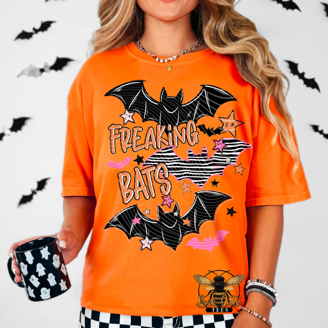 A woman wearing a bright orange graphic t-shirt with a 'FREAKING BATS' design featuring black bats, pink stars, and white text. She is holding a black mug with white ghost patterns, set against a white background decorated with black bat silhouettes.
