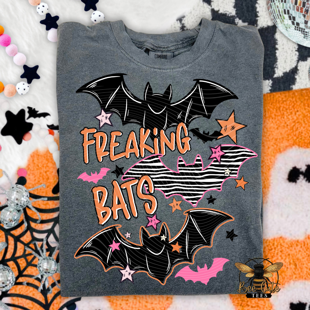 A folded heather gray graphic T-shirt with a Halloween design featuring two black bats, pink and orange stars, and the words 'FREAKING BATS' in orange. The shirt is laid flat on a patterned fabric background with Halloween-themed props.