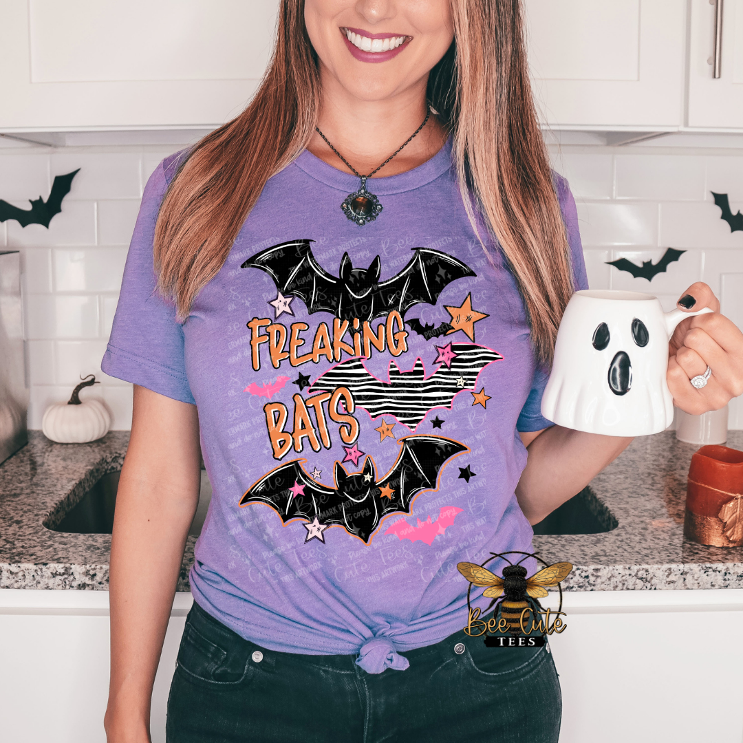 A woman wearing a purple graphic t-shirt with 'FREAKING BATS' text and bat illustrations, holding a white ghost-shaped mug in a kitchen setting.