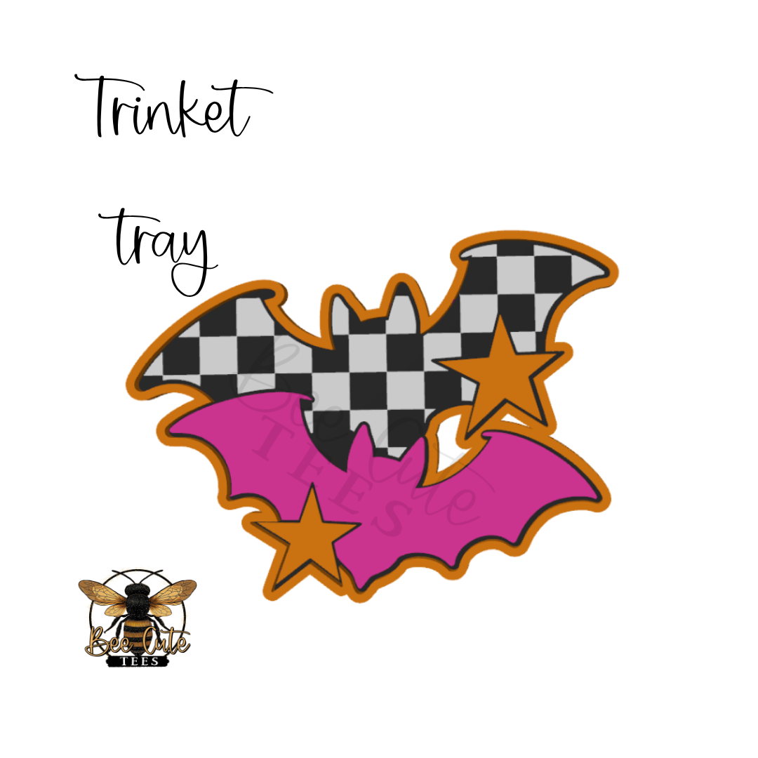 A bat-shaped decorative tray design with black-and-white checkered wings, bright pink wings, orange outlines, and two orange stars. The words 'Trinket tray' appear above, and a small bee logo with 'BEE' is at the lower left on a white background.