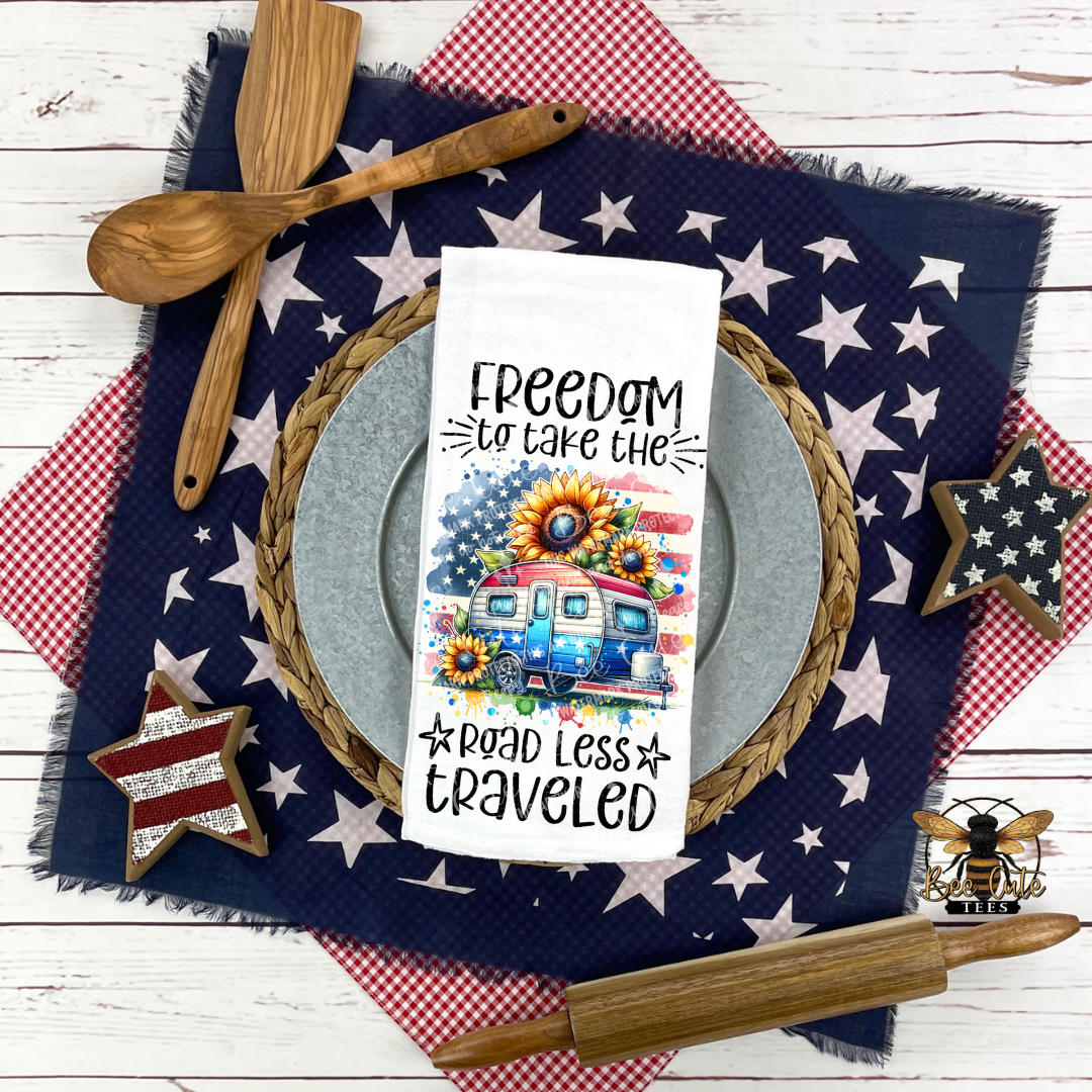 White kitchen towel displayed on a place setting with patriotic decor, featuring a colorful camper with sunflowers and an American flag design and the phrase “Freedom to take the road less traveled,” surrounded by star-patterned cloths, wooden utensils, and rustic accents.
