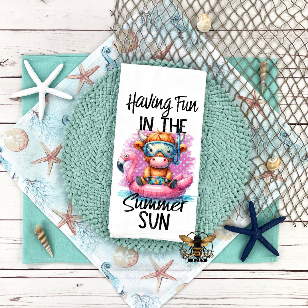 Tea towel with a cow and flamingo design, text 'Having Fun in the Summer Sun', surrounded by beach-themed decor on a wooden surface.