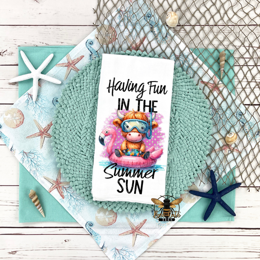 Tea towel with a cow and flamingo design, text 'Having Fun in the Summer Sun', surrounded by beach-themed decor on a wooden surface.