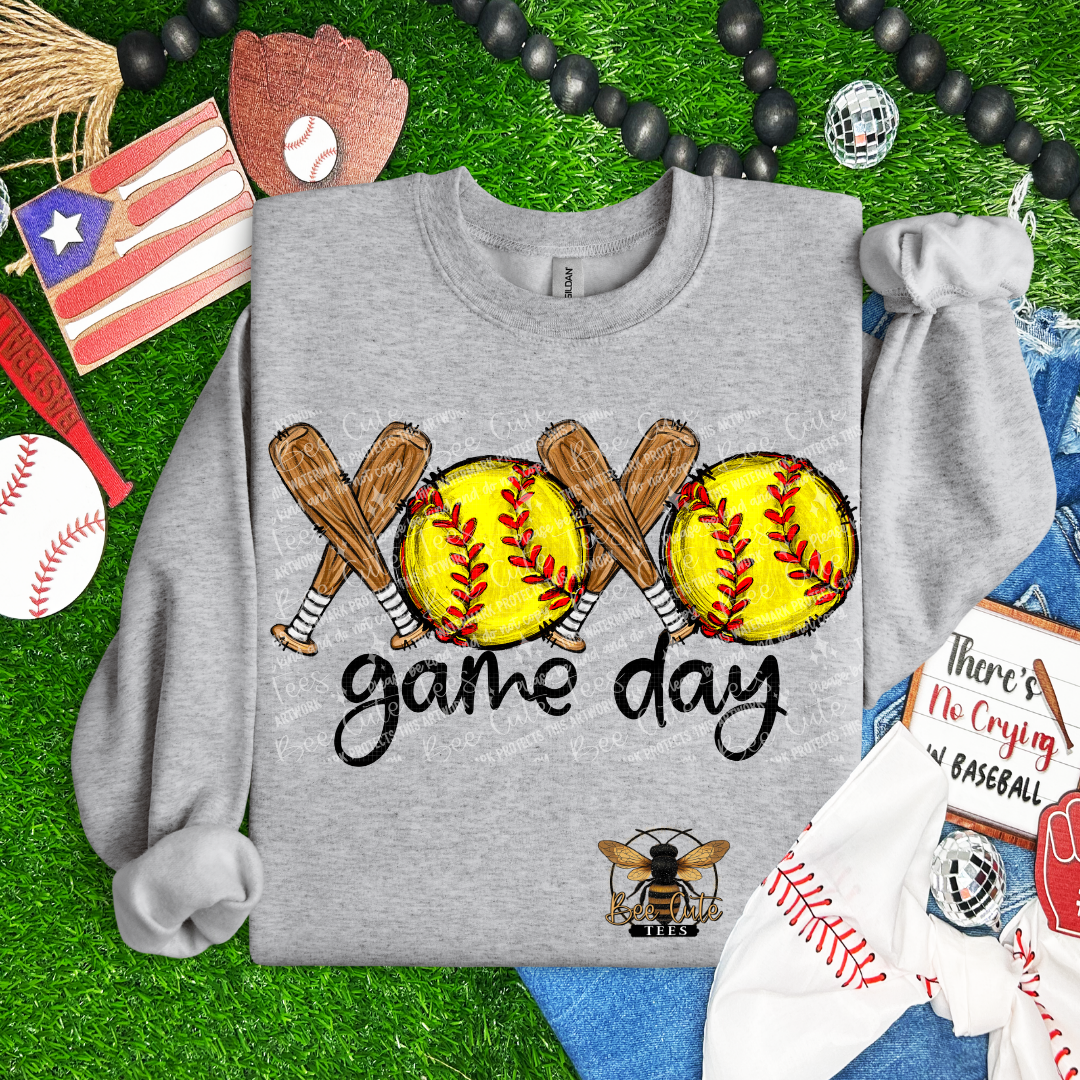 Gray sweatshirt with softball design and 'game day' text on a grass background with sports-themed items.