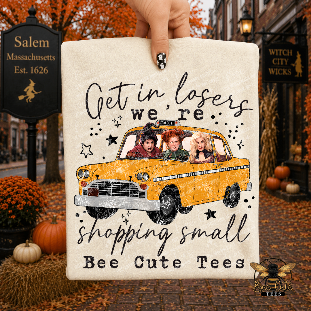 A hand holds a cream-colored folded t-shirt featuring a yellow taxi with the witch characters inside, surrounded by the text 'Get in losers we're shopping small Bee Cute Tees'. The background depicts a cozy, autumn-themed street scene with Salem Massachusetts signs, pumpkins, and a 'Witch City Wicks' brand logo in the bottom right corner.