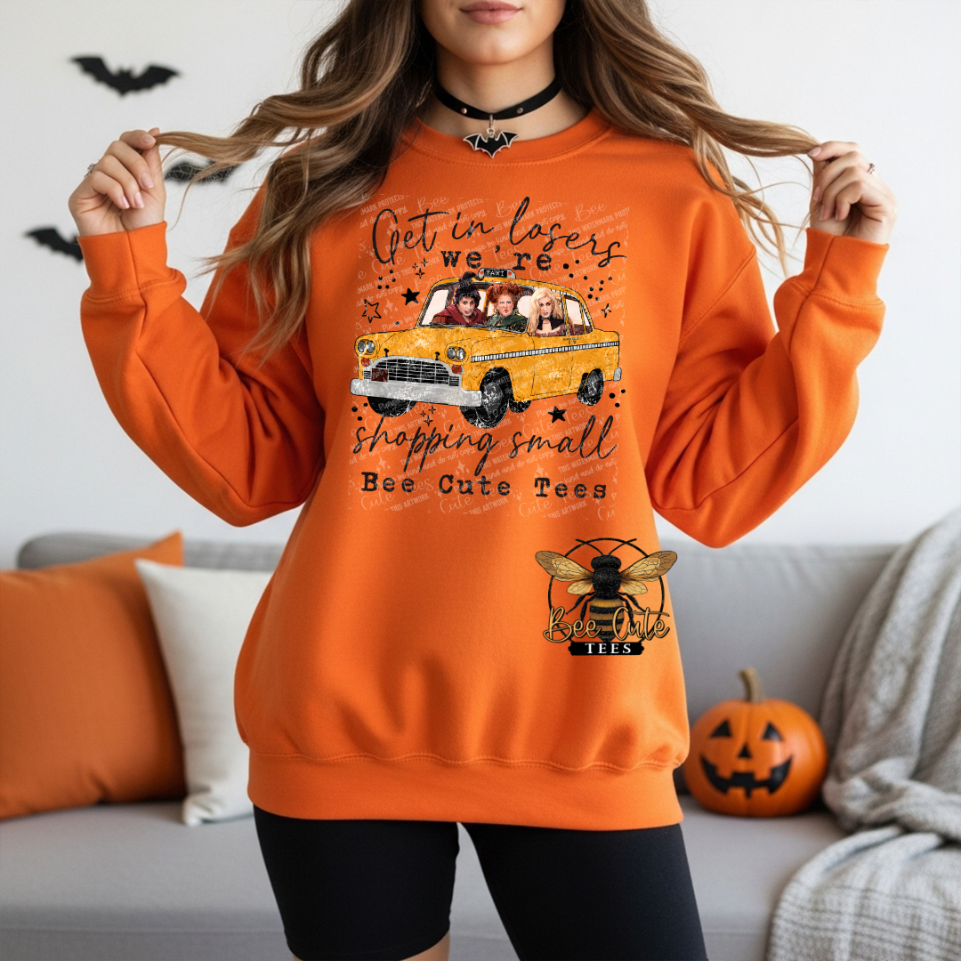 A woman wearing an orange crewneck sweatshirt with a graphic print of a yellow taxi and Halloween-themed text, standing in a living room setting.