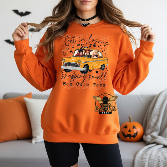 A woman wearing an orange crewneck sweatshirt with a graphic print of a yellow taxi and Halloween-themed text, standing in a living room setting.