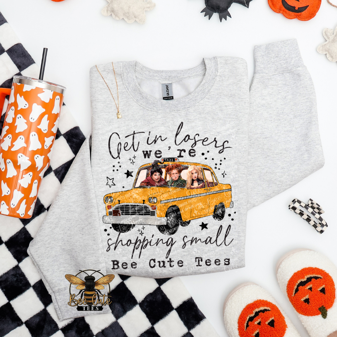 A light heather gray crewneck sweatshirt laid flat on a white surface with Halloween-themed props. The sweatshirt features a graphic of a yellow taxi with four people inside and the text 'Get in losers we're shopping small Bee Cute Tees'.