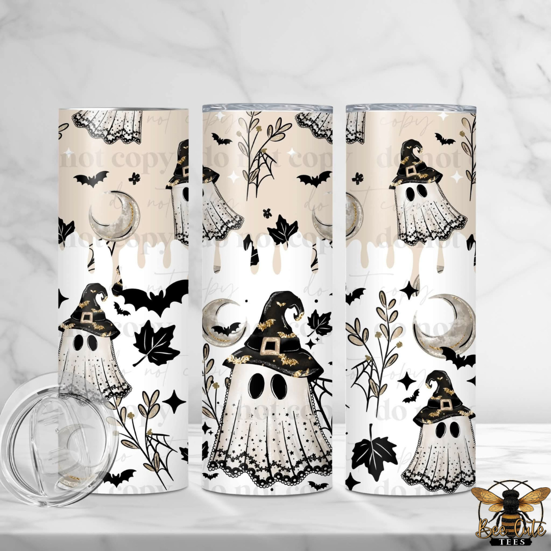 Three tall skinny tumblers with a Halloween-themed print featuring ghosts, witches, bats, and moons on a cream and white background, shown on a light marble surface with a clear lid in the foreground.