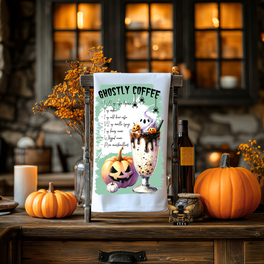 A white kitchen towel with a 'GHOSTLY COFFEE' recipe and cartoon graphics of a ghost and pumpkin, displayed on a wooden stand in a cozy, warmly lit Halloween-themed kitchen setting with pumpkins and candles.