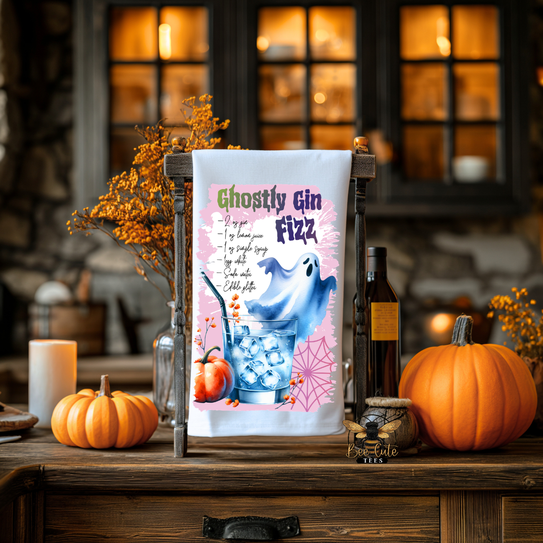A Halloween-themed tea towel with a 'Ghostly Gin Fizz' recipe and illustrations of a ghost, pumpkin, and ice glass, hanging on a wooden stand in a rustic, warmly lit autumnal setting.