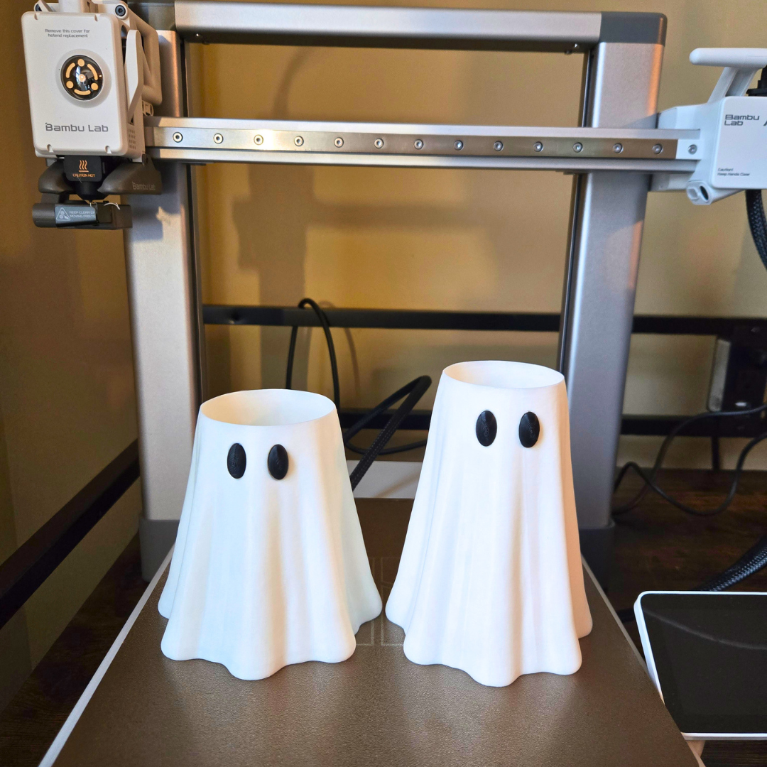 Two white ghost-shaped can sleeves with black eyes printed on a 3D printer bed, with the printer frame visible in the background.