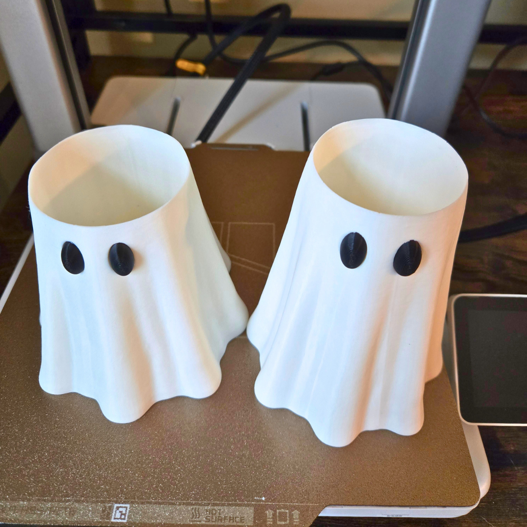 Two white ghost-shaped can holders with black eyes, displayed side-by-side on a brown 3D printer bed.