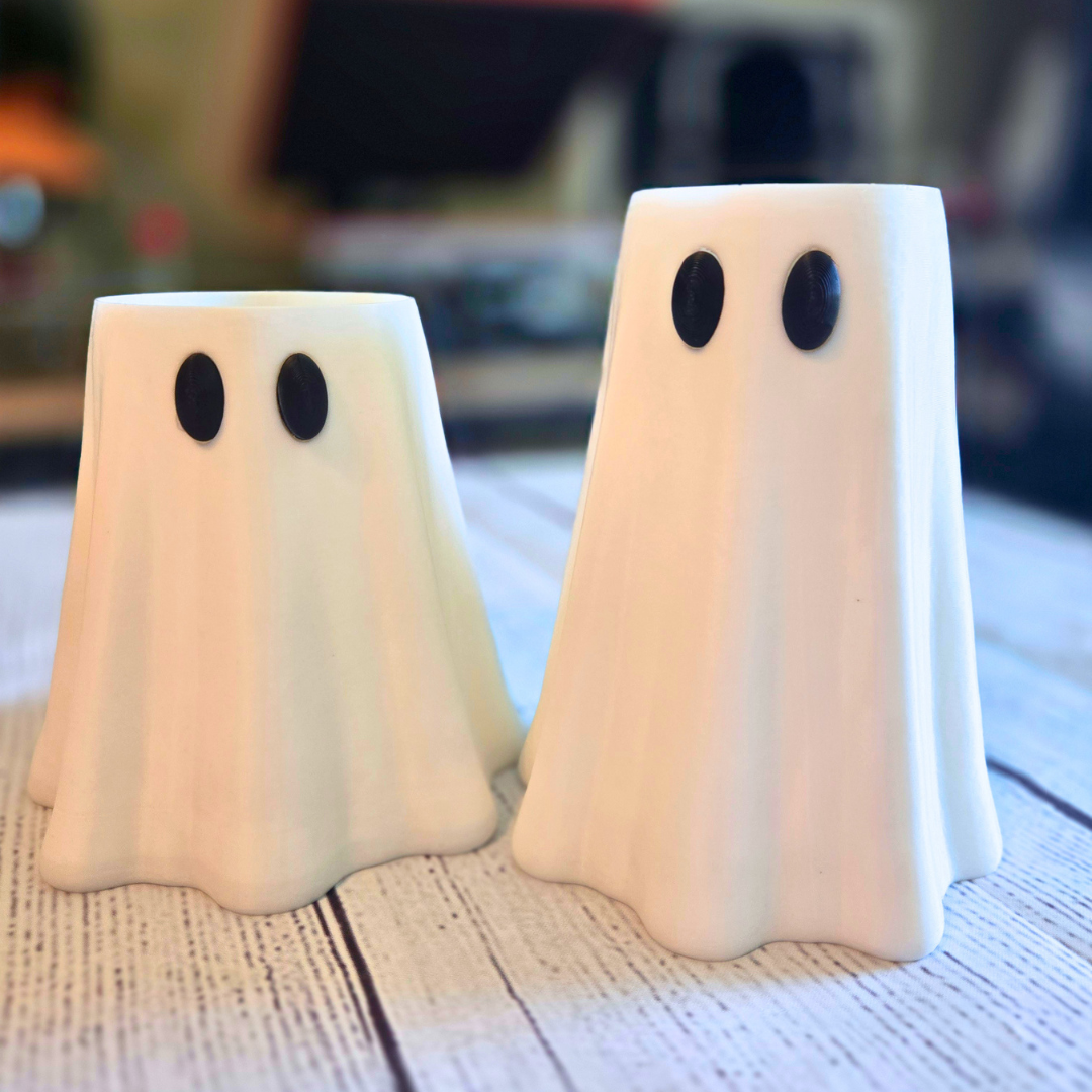 Two ghost-shaped can sleeves with black oval eyes, standing on a rustic white wooden surface with a blurred indoor background.