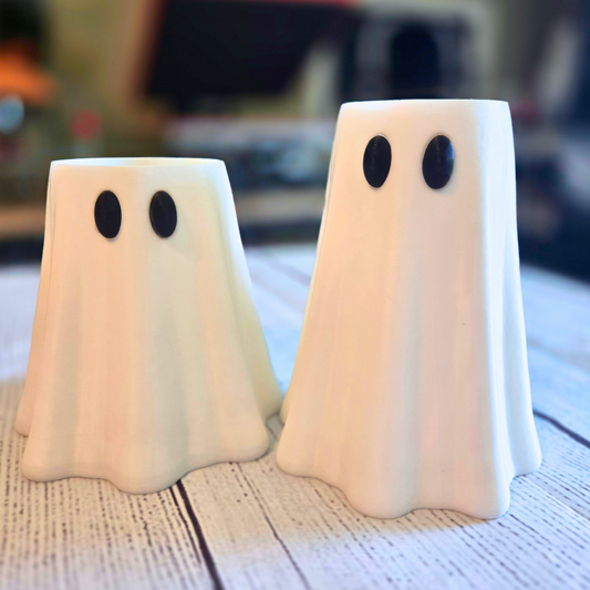Two ghost-shaped can sleeves with black oval eyes, standing on a rustic white wooden surface with a blurred indoor background.