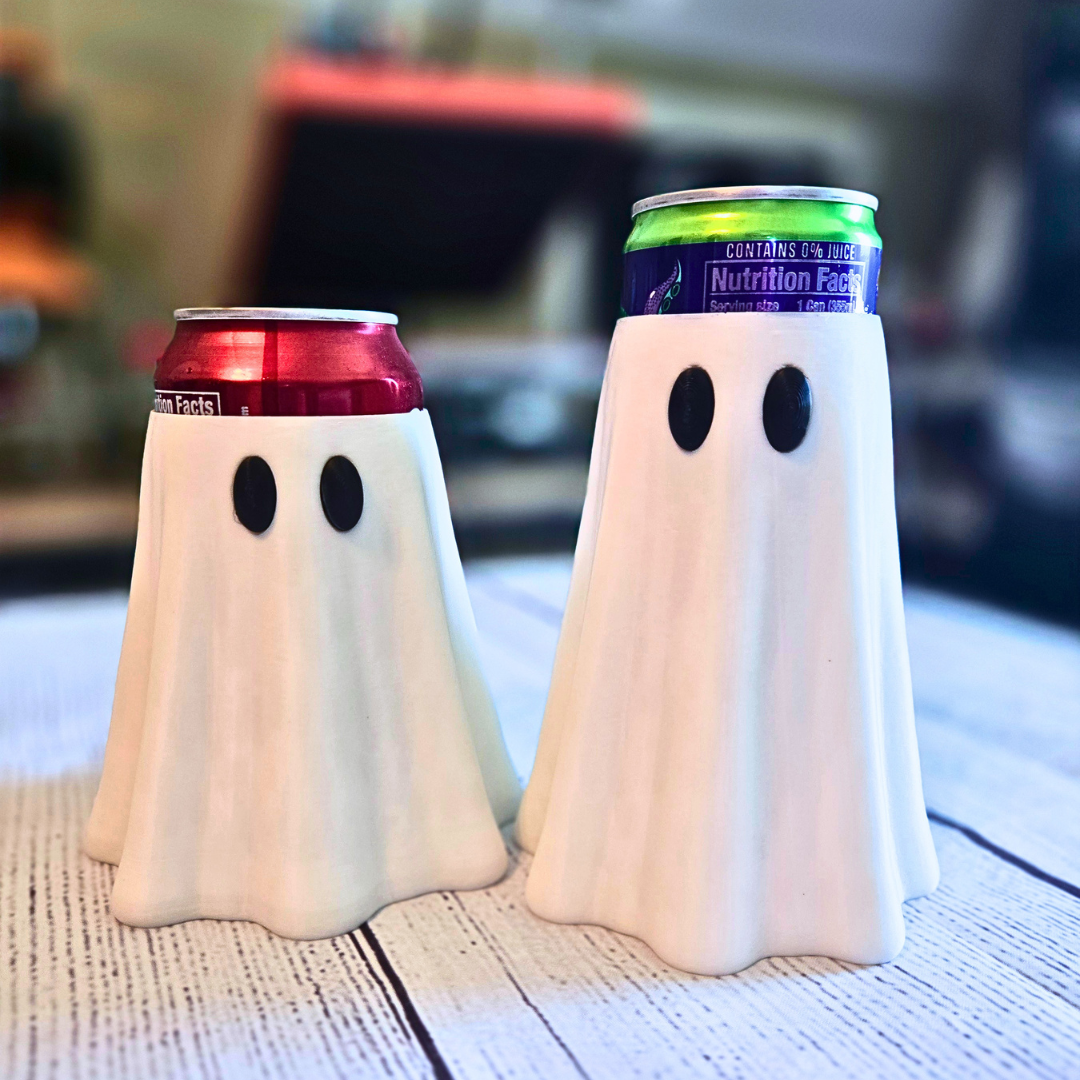 Two ghost-shaped 3D printed can holders on a white wooden table, each holding a beverage can. The left holder contains a red can, and the right holder contains a green can.