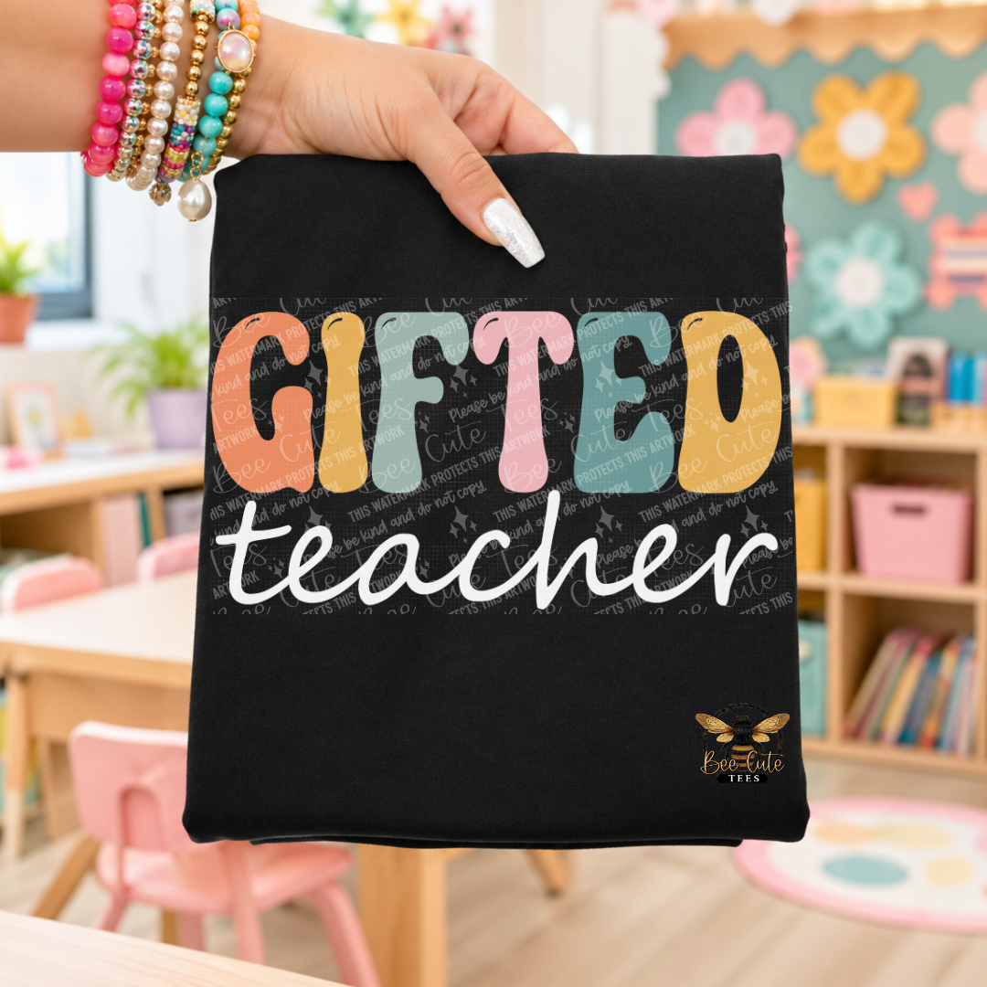A hand wearing colorful beaded bracelets holds up a black t-shirt featuring a colorful graphic that reads 'GIFTED teacher' with a small bee logo reading 'Bee Cute Tees' in the bottom right corner. The background shows a blurred classroom setting.