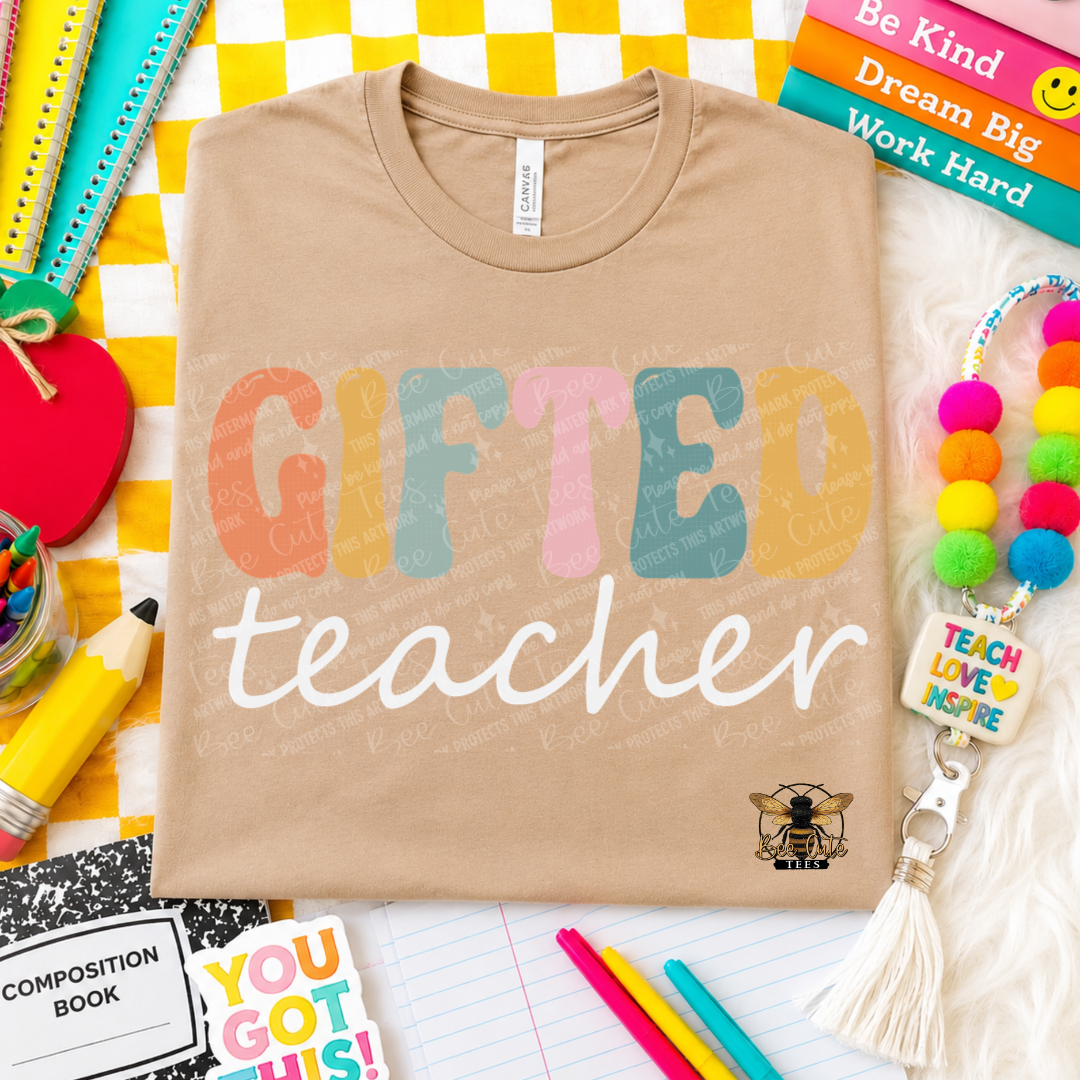 A beige graphic t-shirt laid flat on a white faux fur surface, surrounded by colorful school supplies and books. The shirt features the text 'GIFTED teacher' in a retro, multi-colored bubbly font, with a small bee logo.