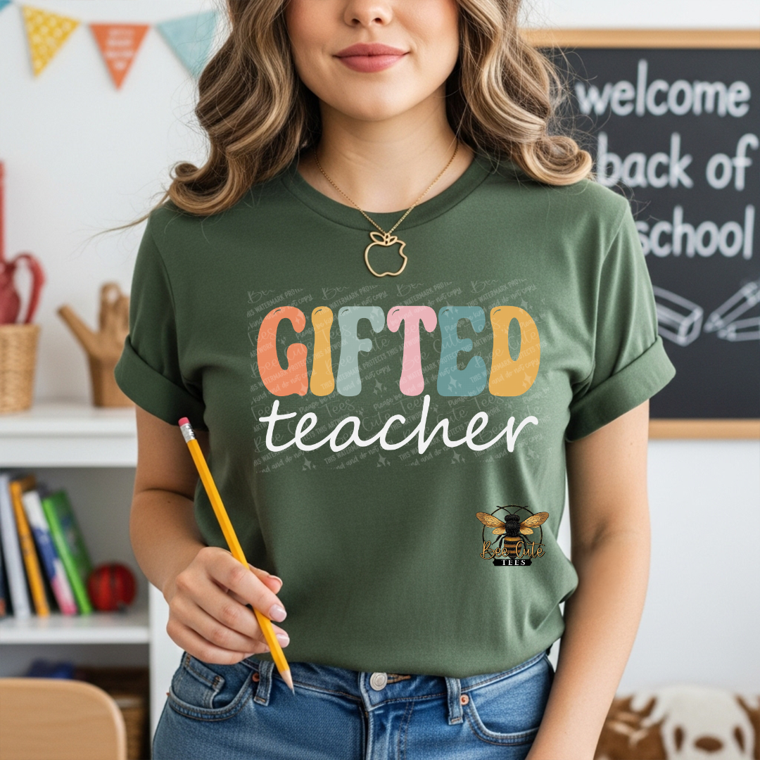 A woman wearing an olive green graphic t-shirt with 'GIFTED teacher' printed in colorful retro letters, holding a yellow pencil in a classroom setting.