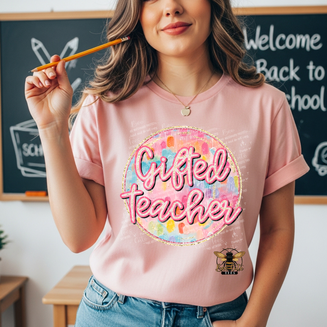 A woman wearing a light pink graphic t-shirt with a colorful circular 'Gifted Teacher' design and a bee logo, standing in front of a chalkboard.