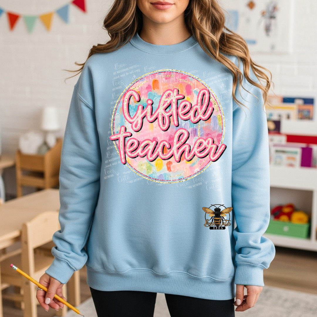 A woman wearing a light blue crewneck sweatshirt with a colorful 'Gifted Teacher' graphic and a bee logo on the front, set in a classroom environment.