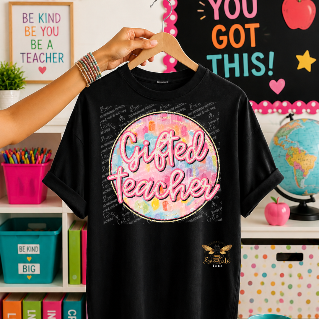 A black graphic t-shirt held on a wooden hanger by a person's hand, featuring a colorful circular 'Gifted Teacher' print on the front, set against a decorated classroom background.