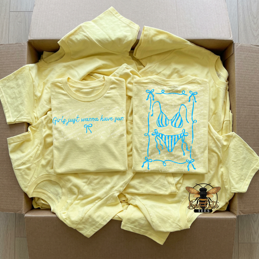 Yellow t-shirts with blue graphics and text in a cardboard box on a wooden floor.