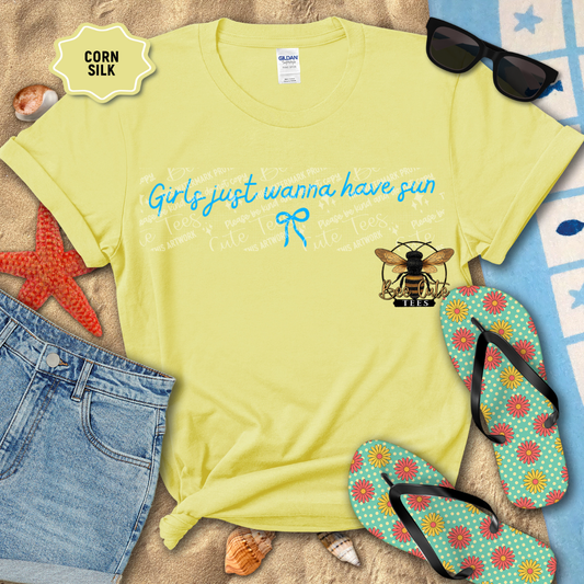Yellow t-shirt with 'Girls just wanna have sun' text and bee logo, surrounded by beach accessories on sand.