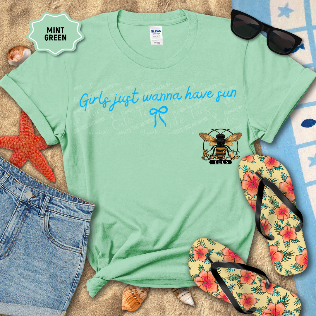 Mint green t-shirt with 'Girls just wanna have sun' text and bee logo, surrounded by beach accessories.