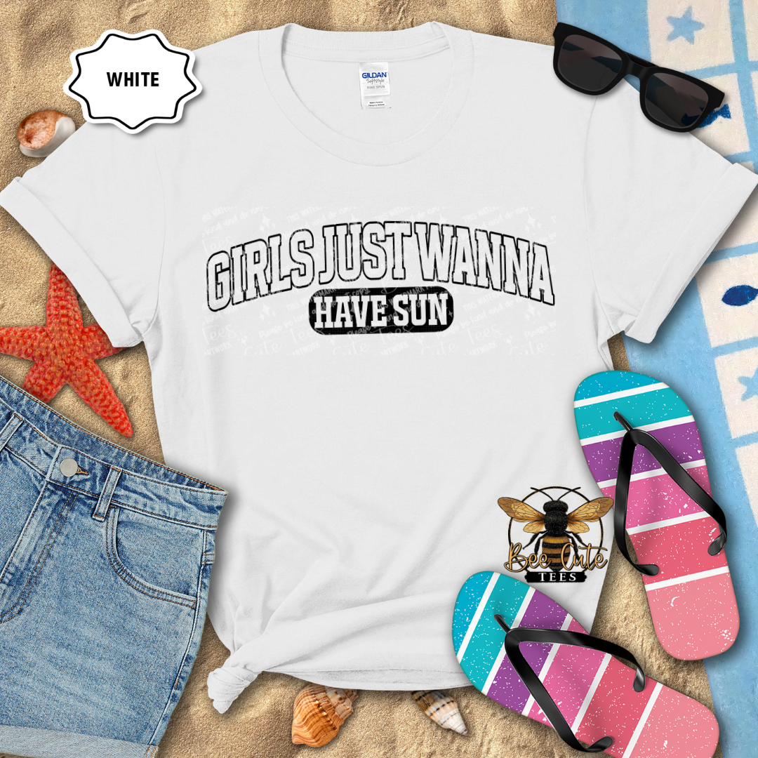 White t-shirt with 'GIRLS JUST WANNA HAVE SUN' text, surrounded by beach-themed items on a sandy background.