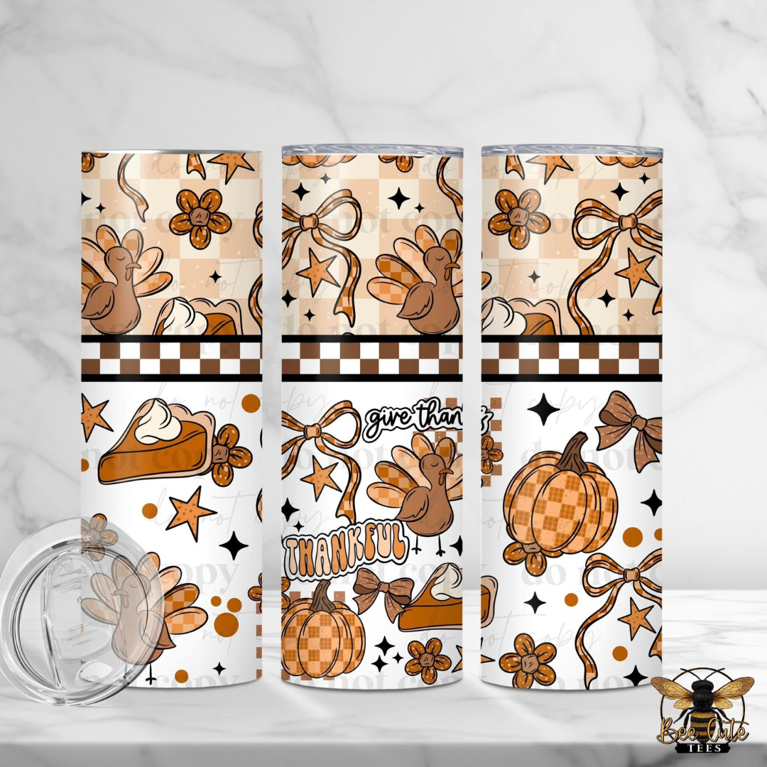 Three tall skinny tumblers with a Thanksgiving-themed autumn pattern, shown on a white marble surface with a clear lid in the foreground. The pattern features pumpkins, turkeys, bows, stars, and the words 'Thankful' and 'give thanks'.