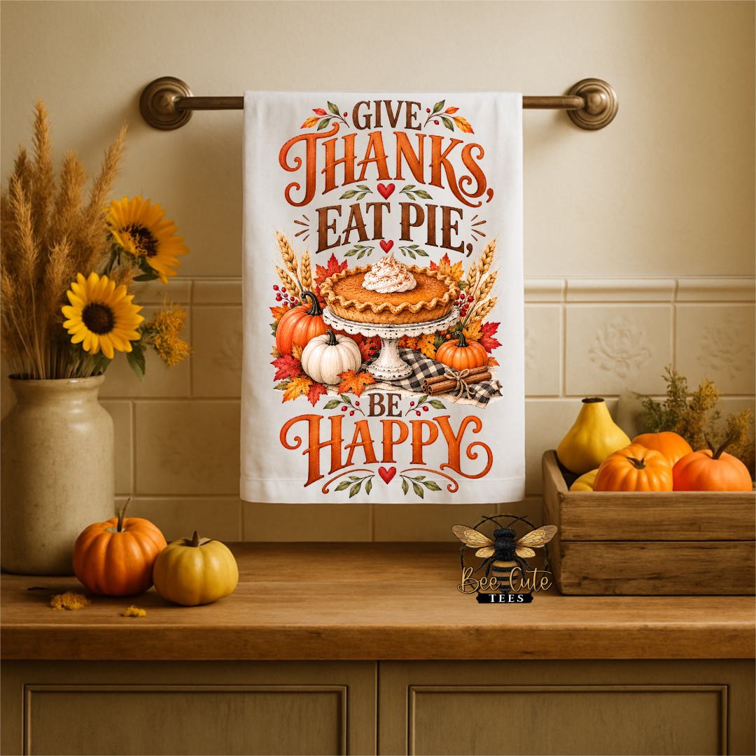 A white kitchen towel with a colorful autumn Thanksgiving design hangs on a brass towel bar. The towel features the text 'GIVE THANKS EAT PIE BE HAPPY' with an illustration of a pie, pumpkins, and fall leaves.