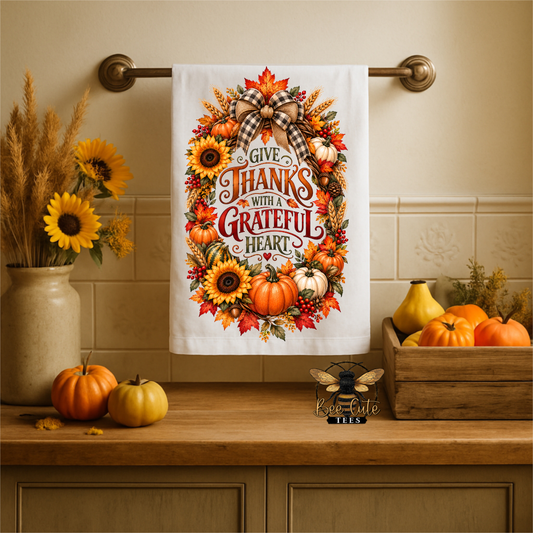 A white kitchen towel with a colorful autumn wreath design and the text 'Give Thanks with a Grateful Heart' hangs on a brass towel bar over a wooden countertop. The scene includes pumpkins, sunflowers, and a decorative bee logo in the foreground.