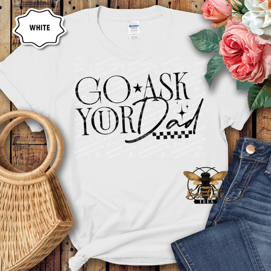 White t-shirt with 'GO ASK YOUR DAD' text, surrounded by flowers and jeans on a wooden surface.