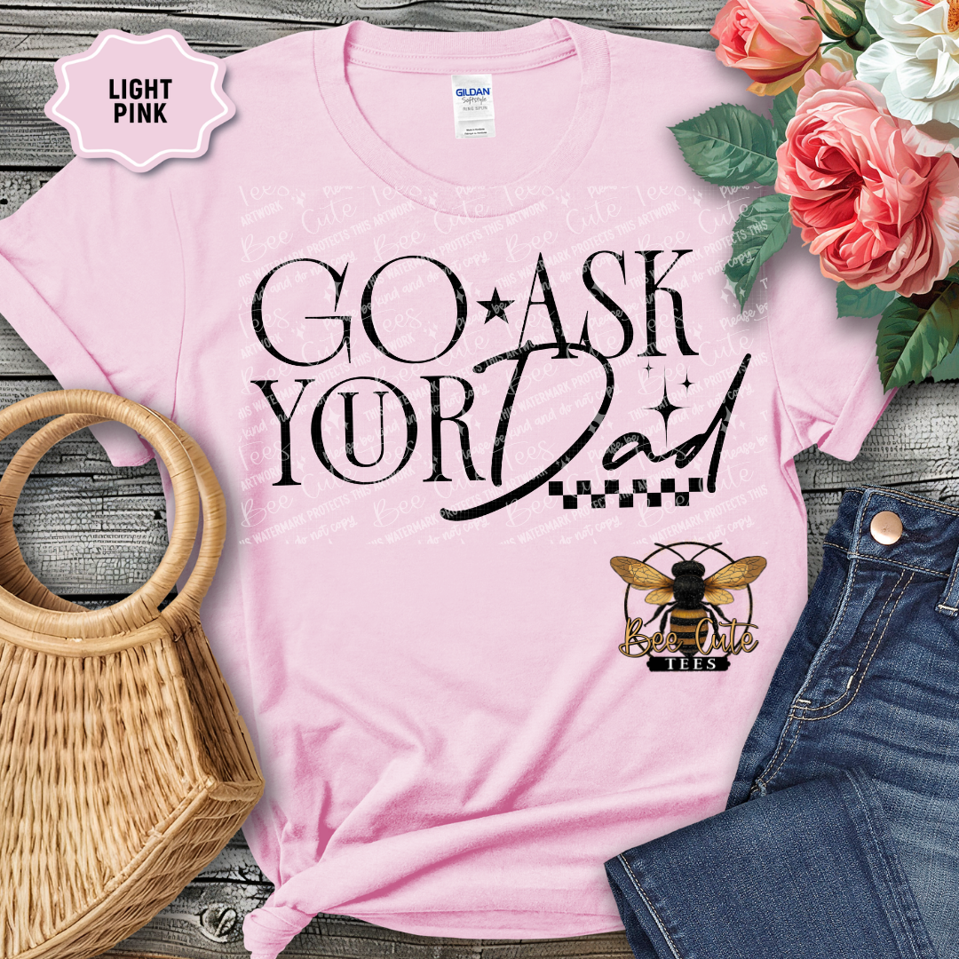Light pink t-shirt with 'GO ASK YOUR DAD' text and bee logo, surrounded by flowers and jeans on a wooden surface.