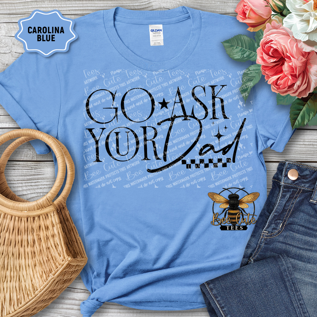 Blue t-shirt with 'GO ASK YOUR DAD' text and bee logo, surrounded by flowers and jeans on a wooden surface.