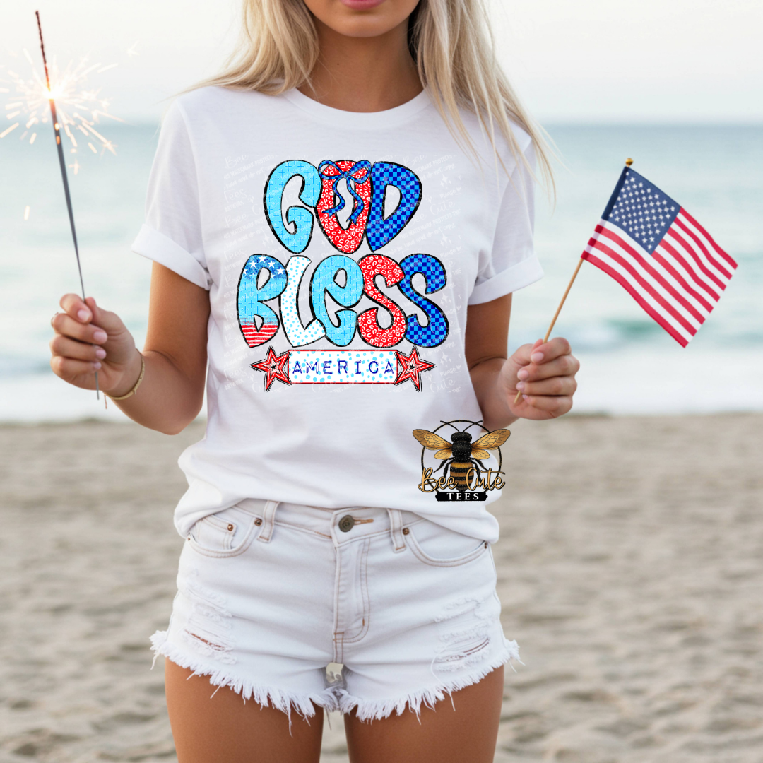 Person wearing a 'God Bless America' t-shirt on a beach with an American flag and sparkler.