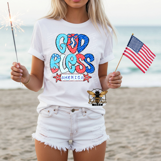 Person wearing a 'God Bless America' t-shirt on a beach with an American flag and sparkler.