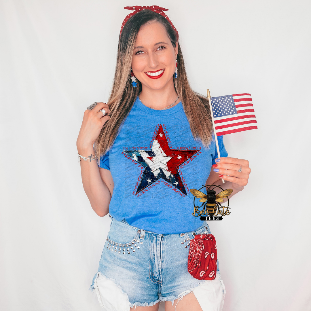 Woman wearing a heather royal blue patriotic graphic tee featuring a distressed red, white, and blue star design with “God Bless America” text accents, paired with denim shorts and patriotic accessories while holding a small American flag.
