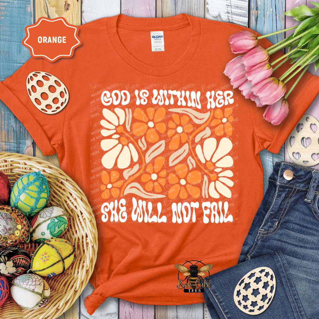 Orange t-shirt with floral design and text 'God is within her, she will not fail' on a wooden surface with Easter decorations.